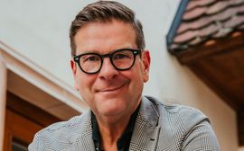 Mann mit Brille im karierten Sakko vor Hotelfassade, freundlich blickend