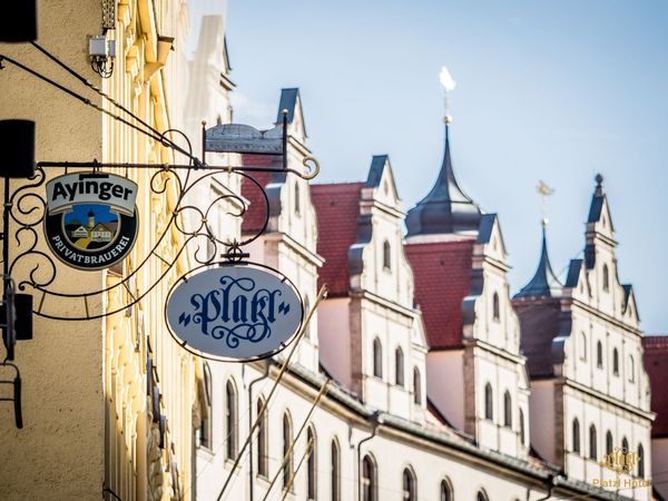 Ein Blick entlang Münchnener Häuserfassaden, an denen neben dem Logo der Brauerei Ayinger auch ein Logo des Platzl Hotels München hängt