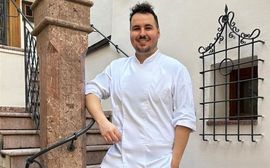 A smiling chef in white uniform poses casually on stone steps beside a historic building