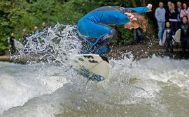 Ein Surfer in blauem Neoprenanzug an der Eisbachwelle vor Zuschauern.