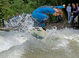 A surfer in blue wetsuit at the Eisbach wave in front of spectators.